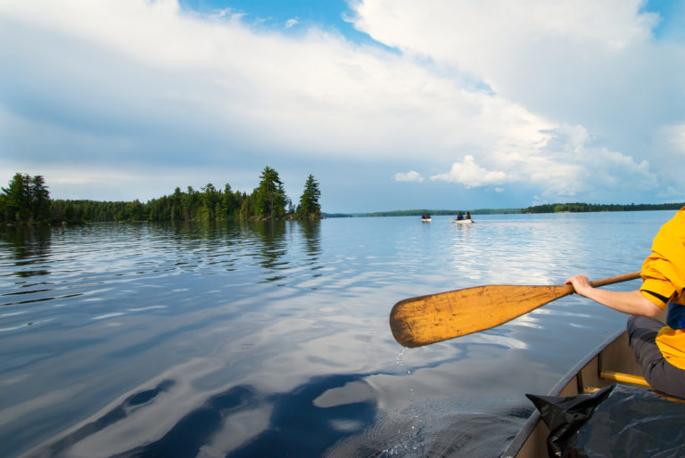 Person paddling a canoe on a calm lake with trees in the background